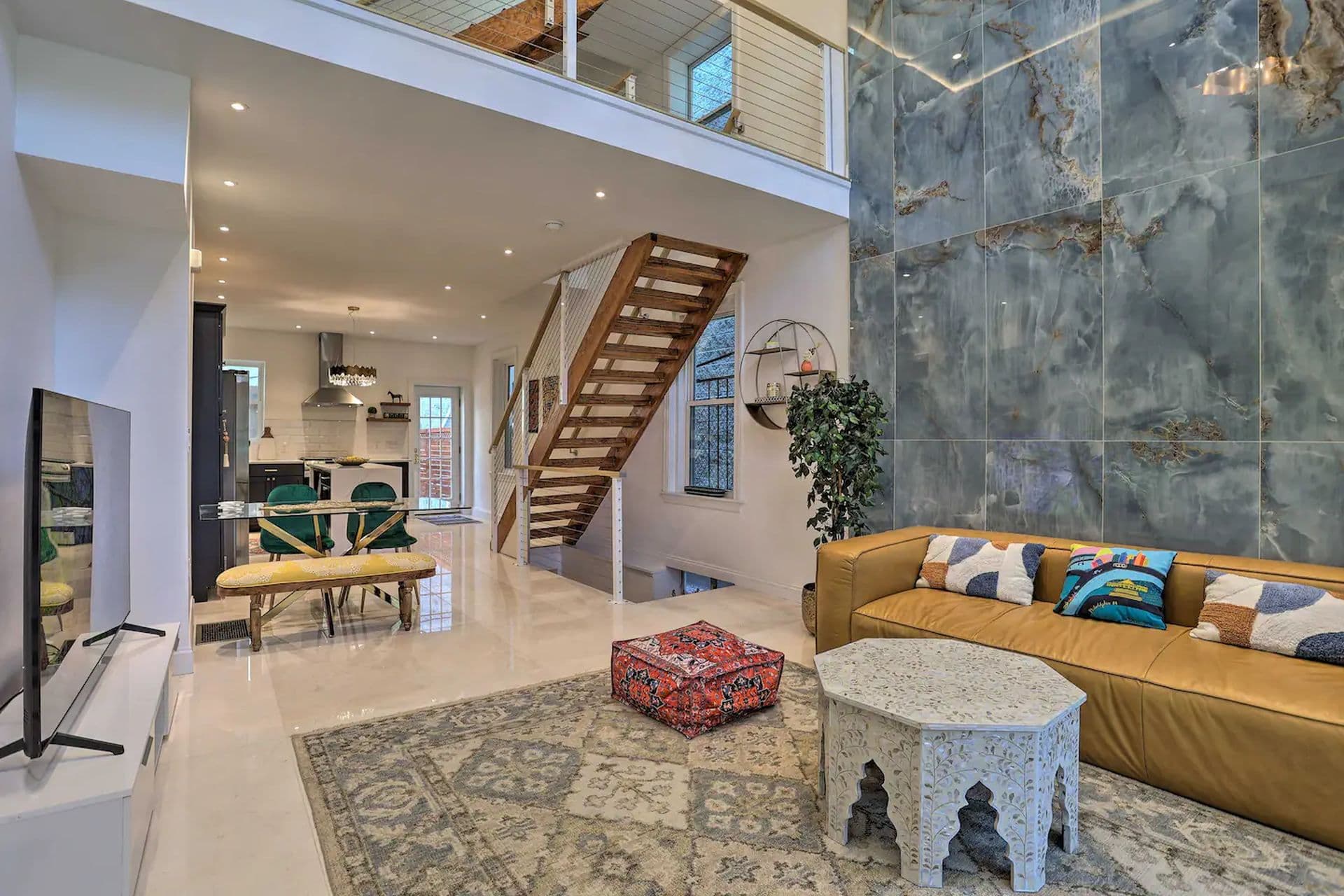 Modern open plan living room with high ceilings and a wooden staircase leading to an upper level. A brown sofa with colorful cushions sits on a patterned rug, facing a small ornate table. A glimpse of the kitchen and dining area is visible in the background.
