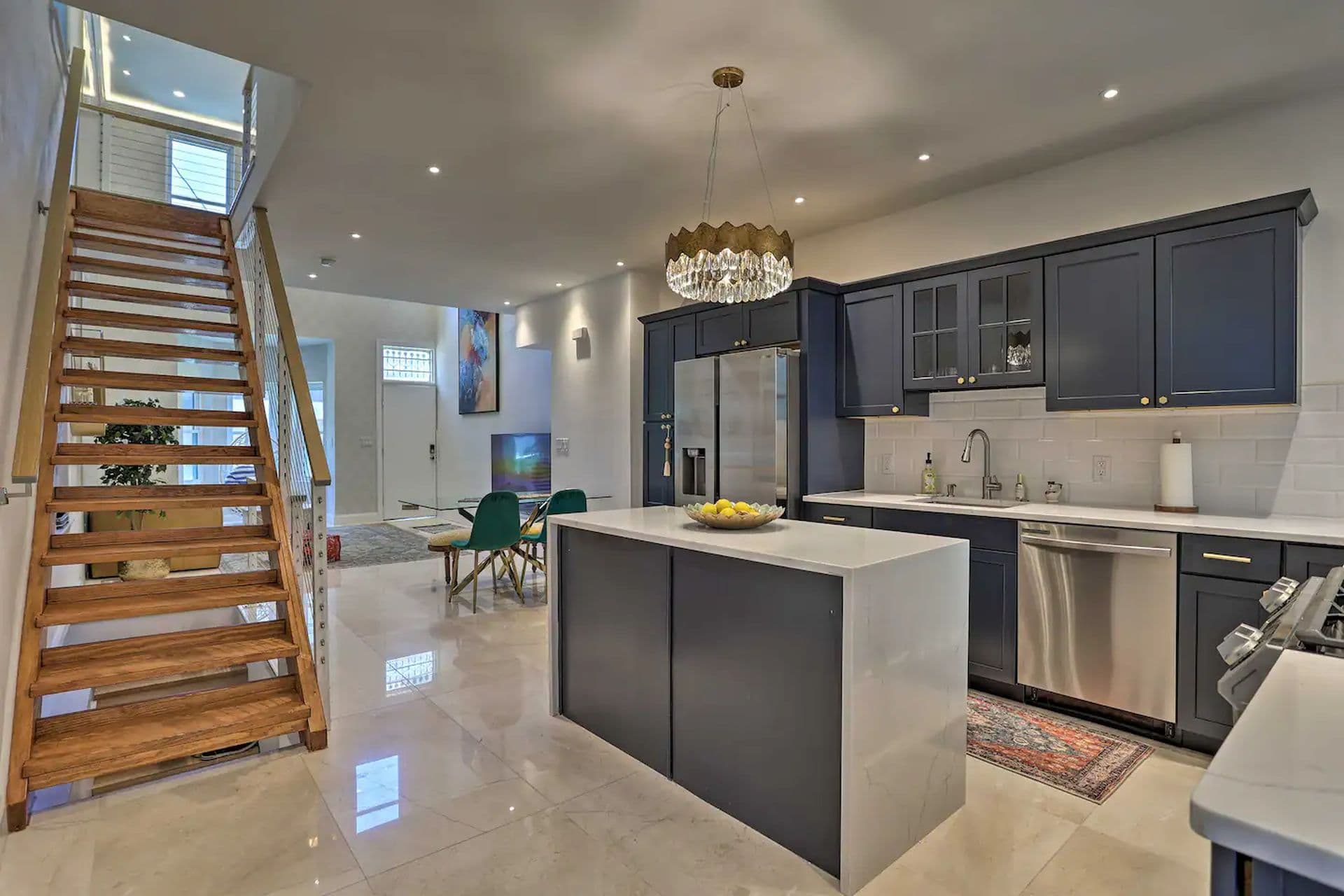 A modern kitchen with dark blue cabinets and a white island countertop. A wooden staircase leads to the second floor. A dining area and living room are visible in the background.