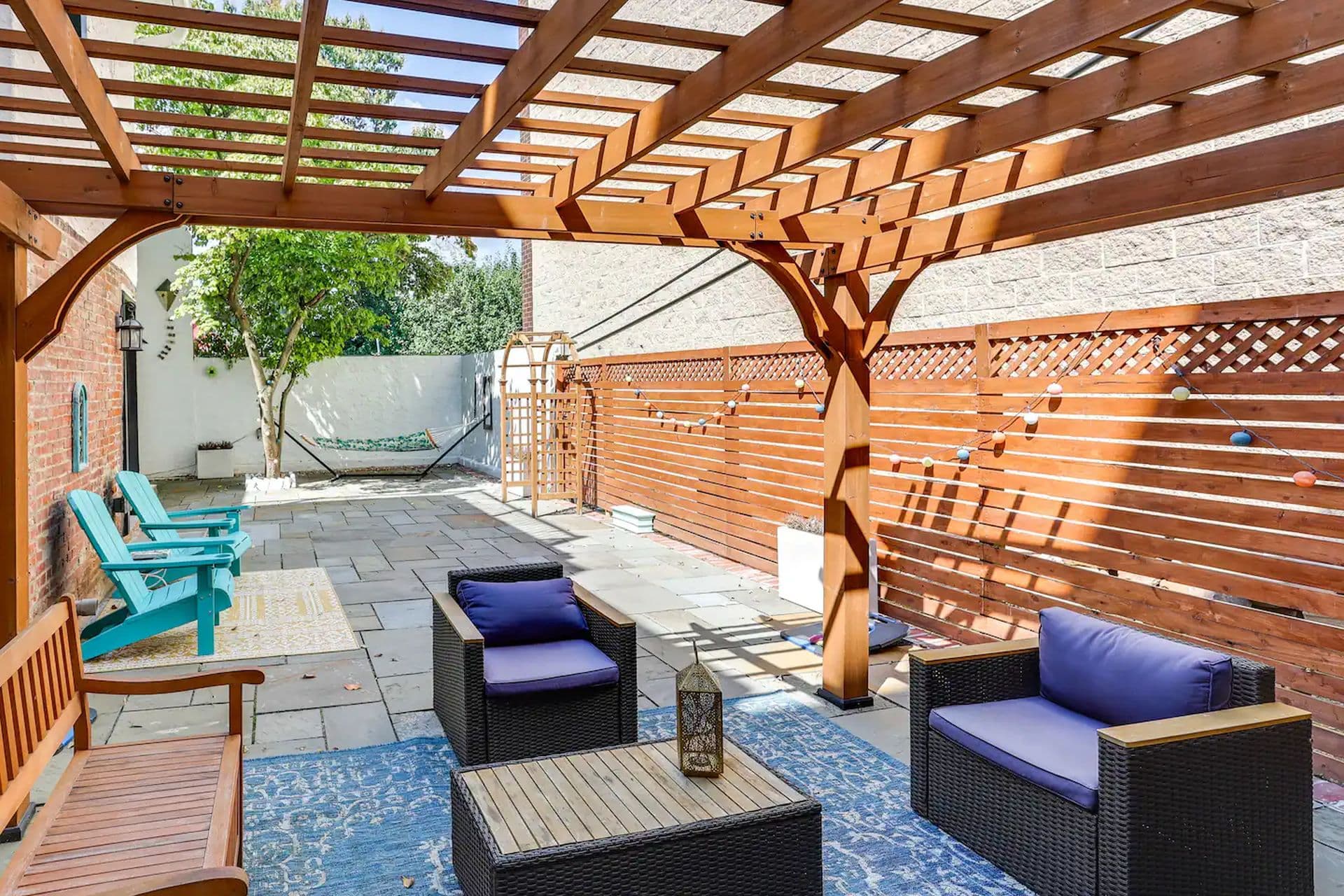 A backyard patio area with a wooden pergola providing shade. Two wicker chairs with blue cushions sit on an outdoor rug, facing a small table. A hammock is strung across the back of the yard.