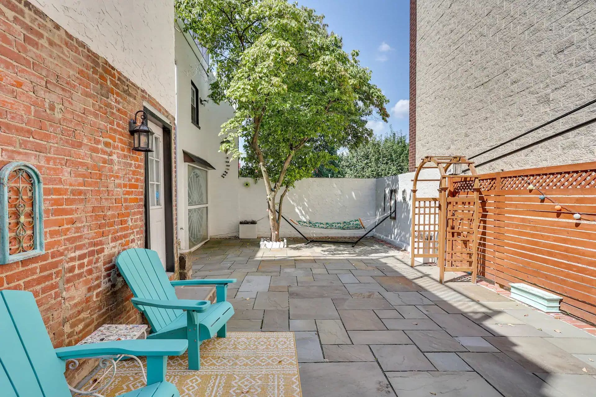 A paved backyard patio with two turquoise Adirondack chairs and a hammock strung between a tree and a brick wall.