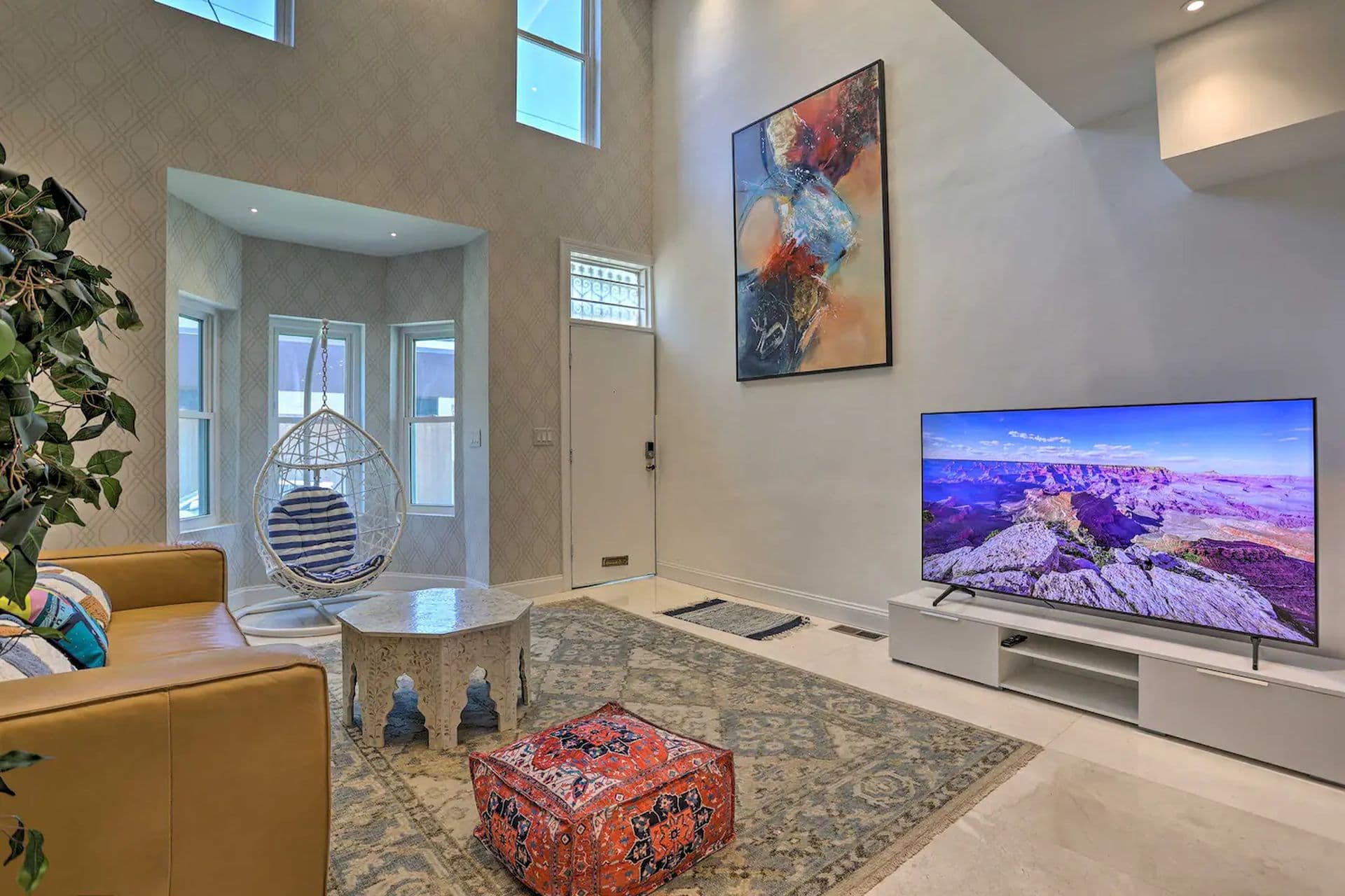 A modern living room with a sunken seating area featuring a brown couch, colorful cushions, and a patterned rug. A large TV mounted on the wall displays a scenic landscape. Large windows and a unique piece of artwork adorn the walls.