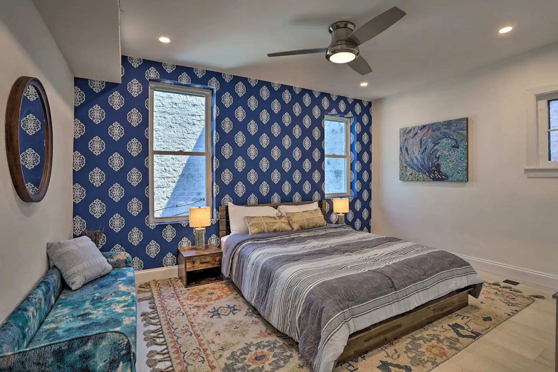 A bedroom with a king-size bed with a striped comforter. The bed is against a wall with blue patterned wallpaper. There is a small bench at the foot of the bed, and a window on either side of the bed.