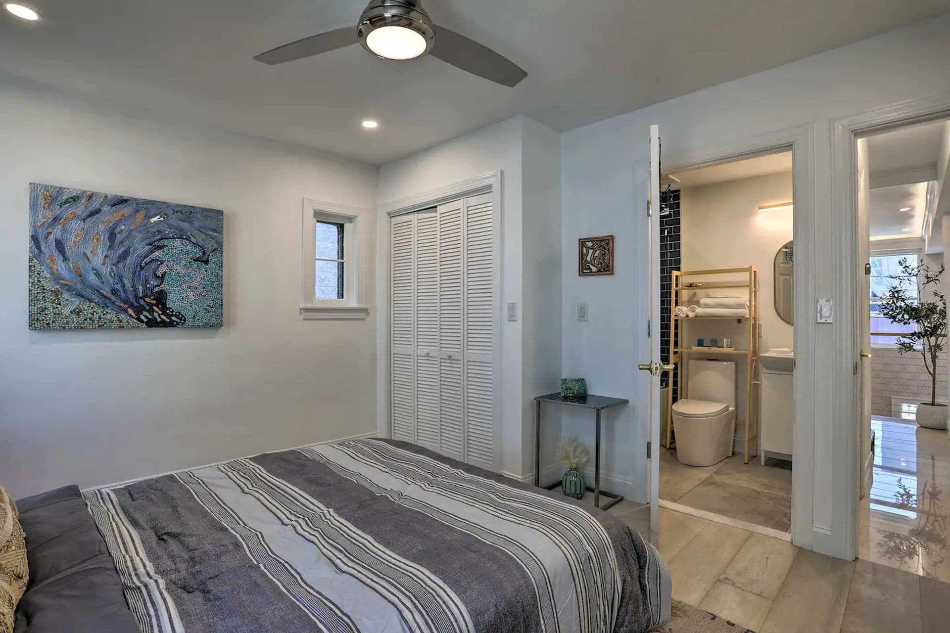 A bright bedroom with a bed with a striped comforter. A bathroom with a glass shower enclosure is visible through the open door. White louvered closet doors are on the left and a window is on the right.