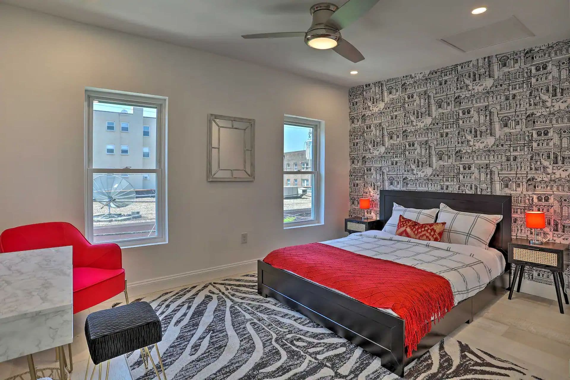 A bedroom with a bed topped with a red and white comforter on a zebra-print rug. One wall has a black and white graphic wallpaper. A red chair sits beside the bed and two windows provide natural light.