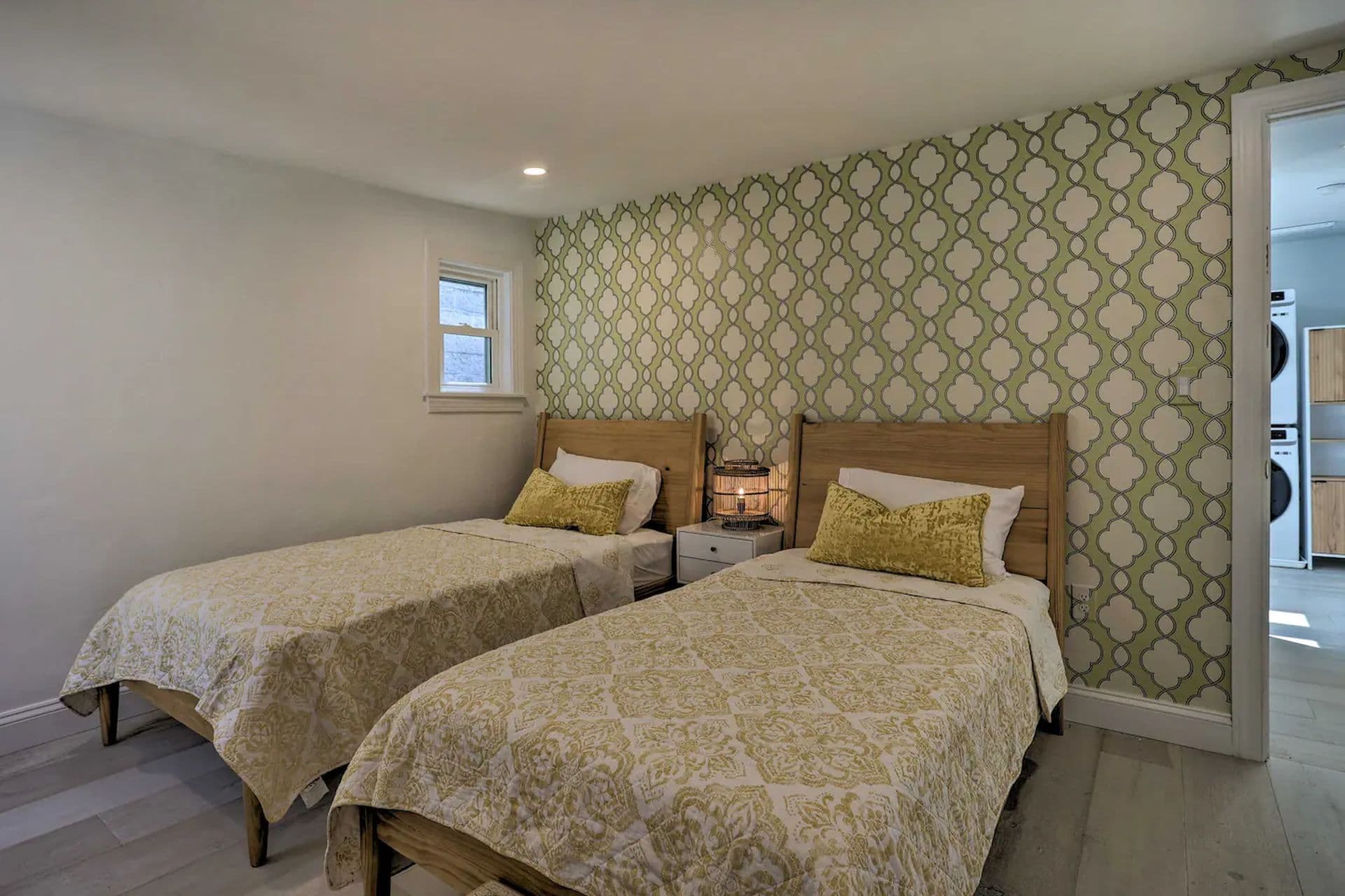 A bedroom with two twin beds with beige patterned comforters. The beds are against a wall with a green geometric patterned wallpaper. A window is on the left wall, and a door to a bathroom is slightly ajar on the right.