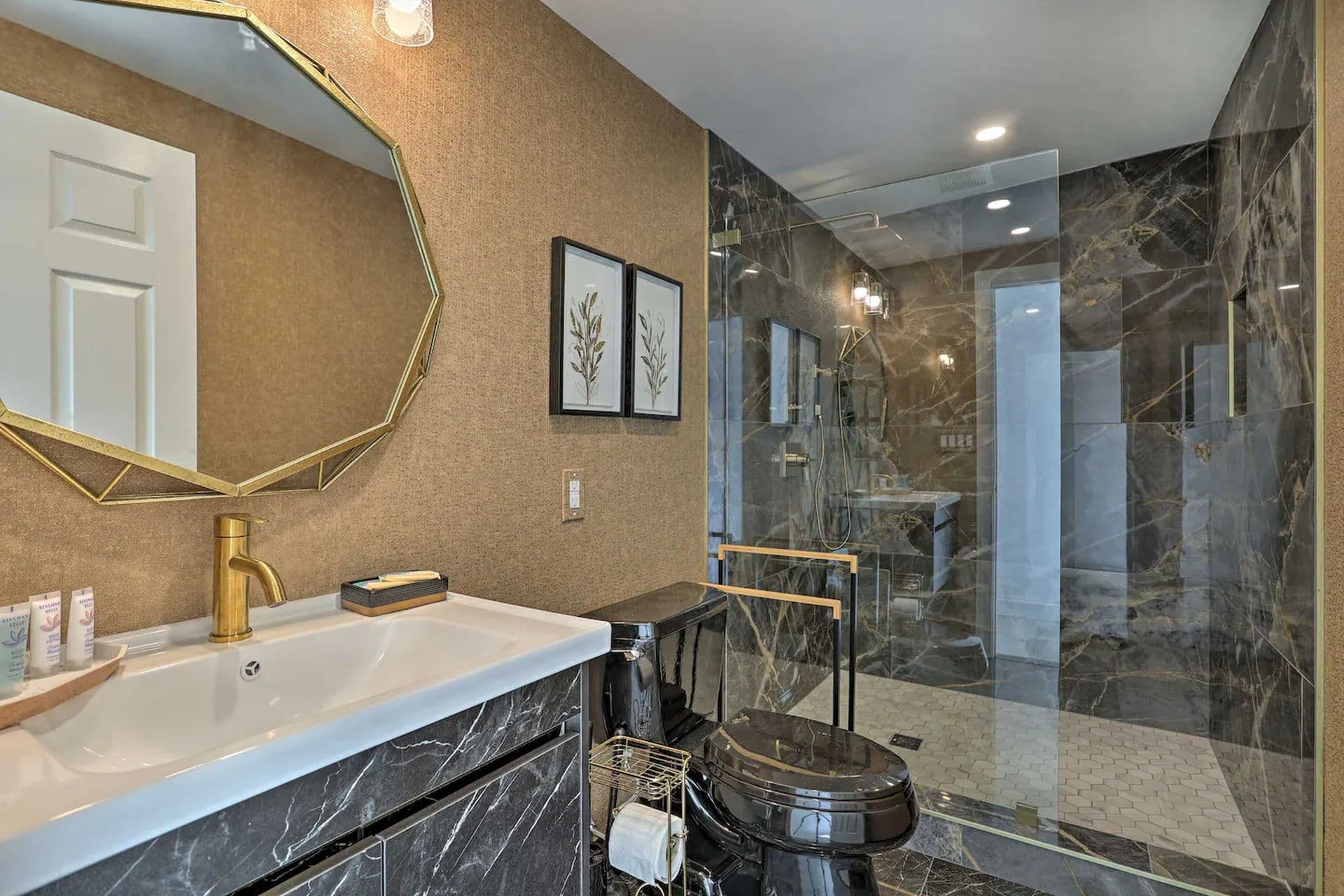A modern bathroom with a large walk-in shower with marbled tile walls. A white vanity with a gold framed octagonal mirror above it sits outside the shower.