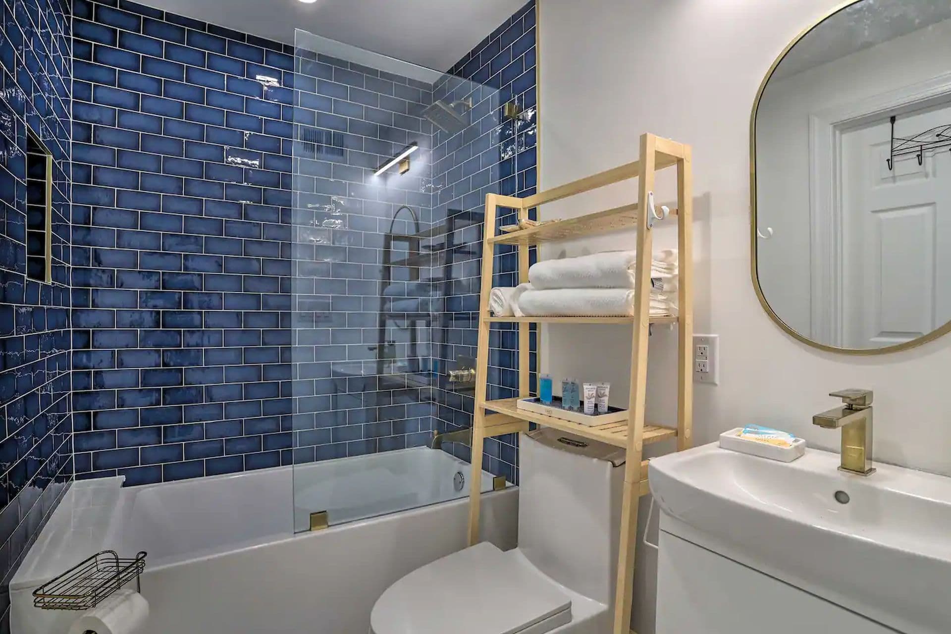 A bathroom with a white sink and toilet, and a bathtub with a blue tiled shower surround. A wooden shelving unit holds towels and toiletries.
