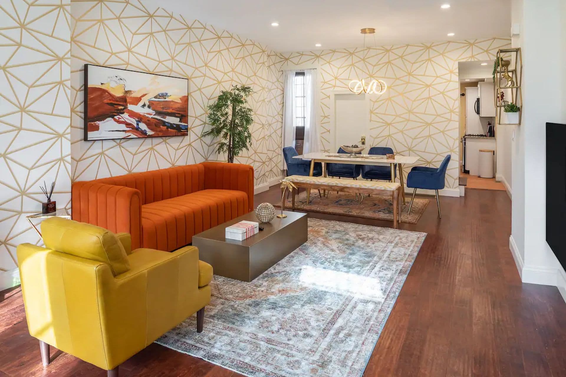 A stylish living room with an orange sofa and a yellow armchair on a patterned rug. A wooden coffee table sits in front of the sofa. In the background, a dining area with a table and chairs is visible.