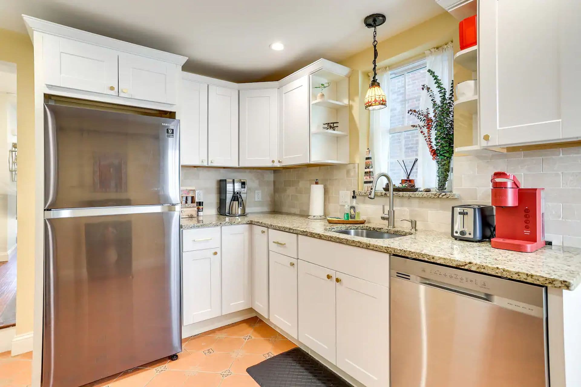 A kitchen with white cabinets and tan countertops. A stainless steel refrigerator is on the left, and a window is above the sink.