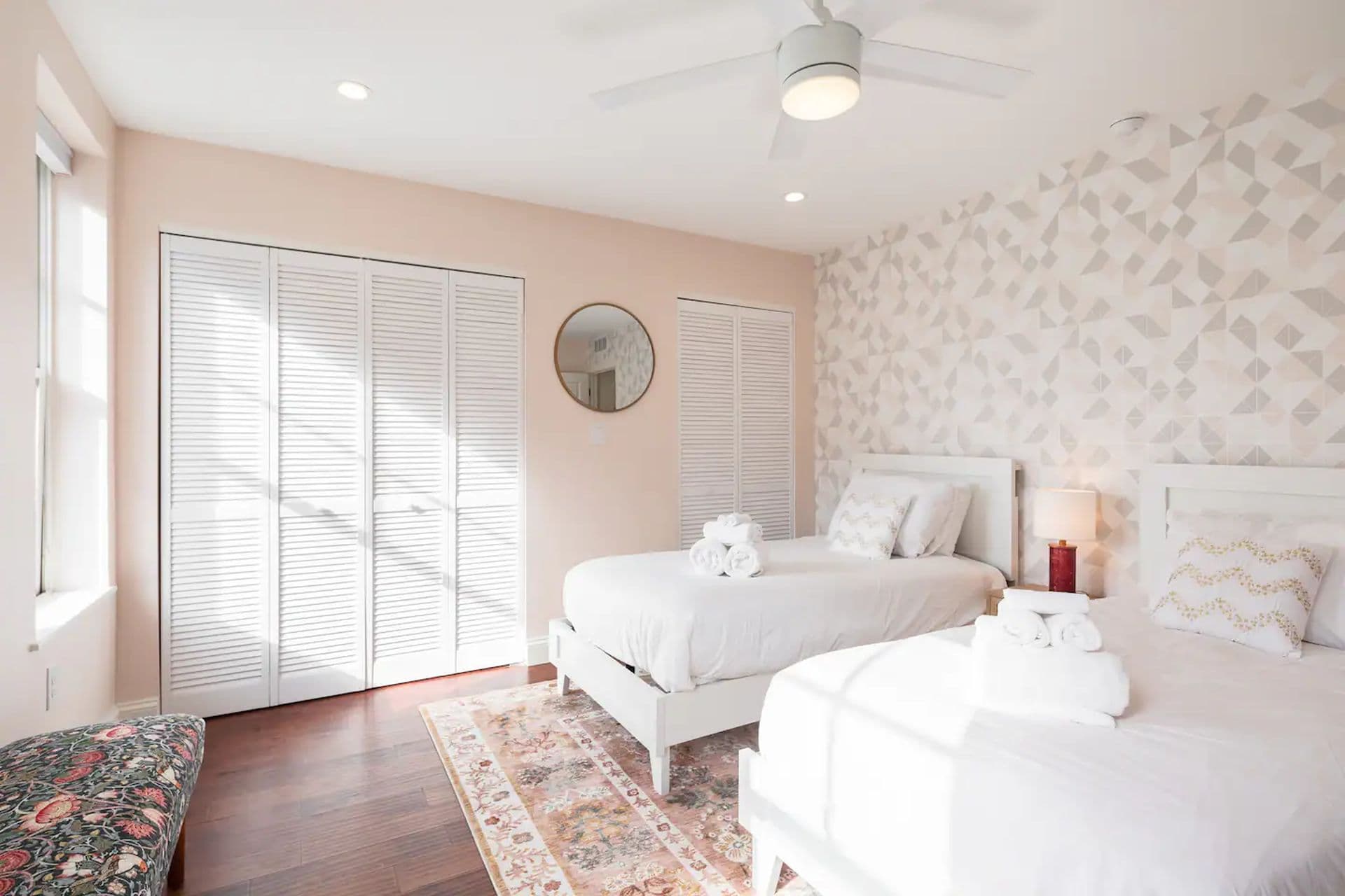 A bedroom with two single beds with white comforters. A floral wallpaper covers one wall, and white closet doors are on another. A window allows natural light to stream in.