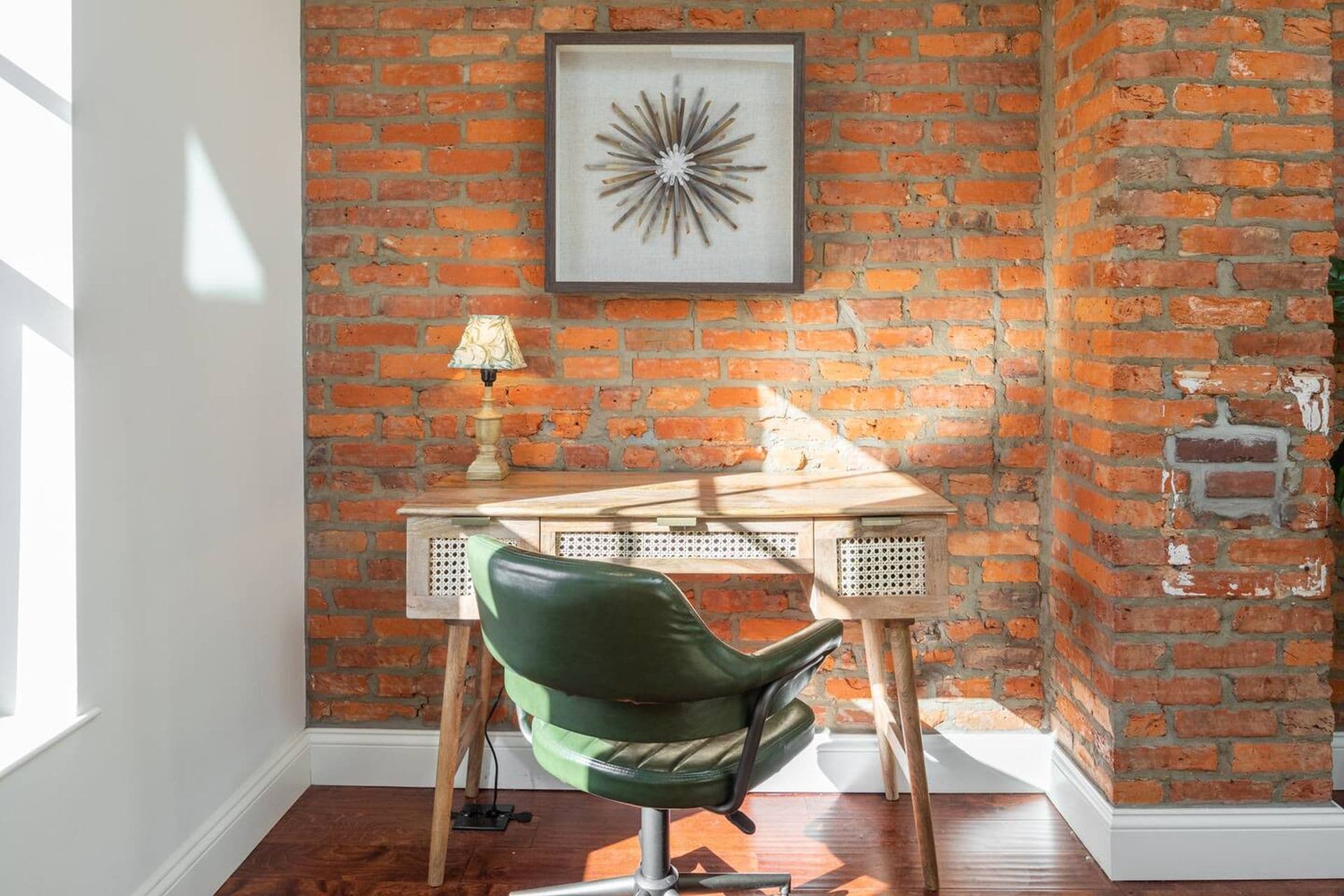 A home office with a wooden desk and a green office chair set against a red brick wall. A framed art piece hangs above the desk, and a small lamp sits on the desk's surface.