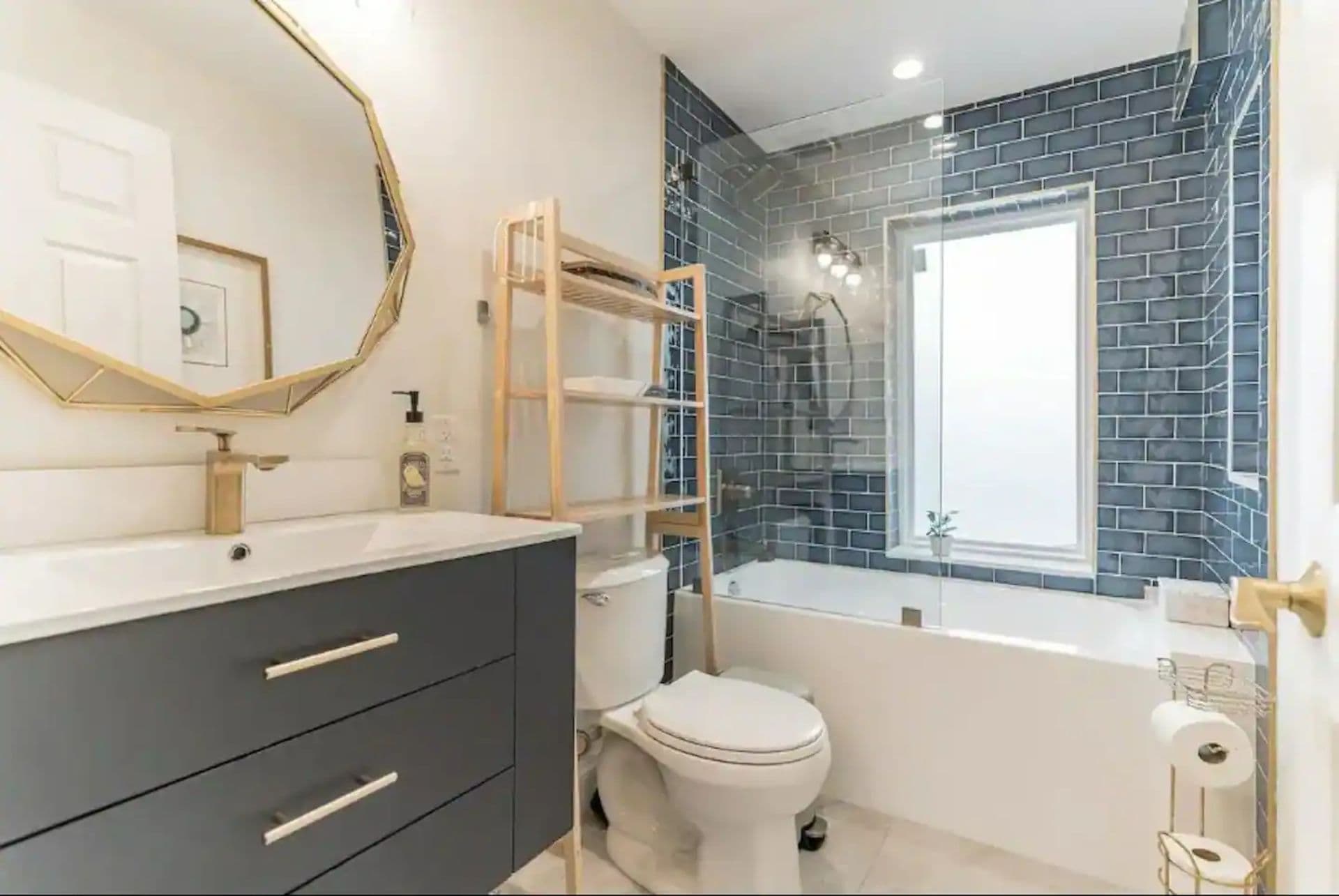 A bathroom with a gray vanity, a white toilet, and a bathtub with a tiled shower surround. A large, geometrically shaped mirror hangs above the vanity, and a wooden shelf unit provides storage for towels.