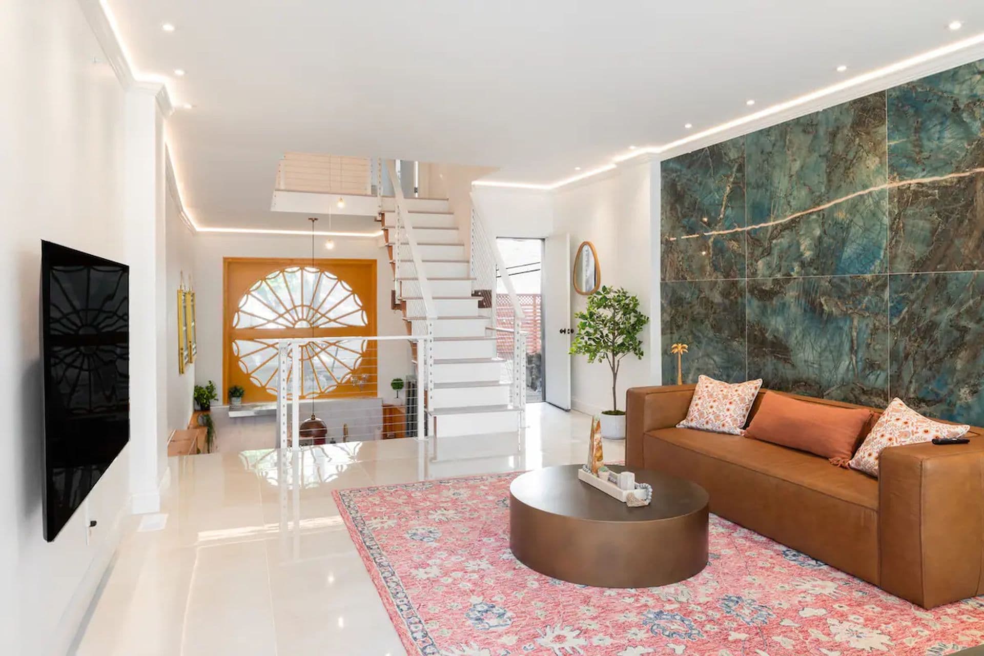 A modern living room with a brown sofa and a round coffee table on a patterned rug. A staircase leads to an upper level, and a large abstract artwork hangs on the wall.