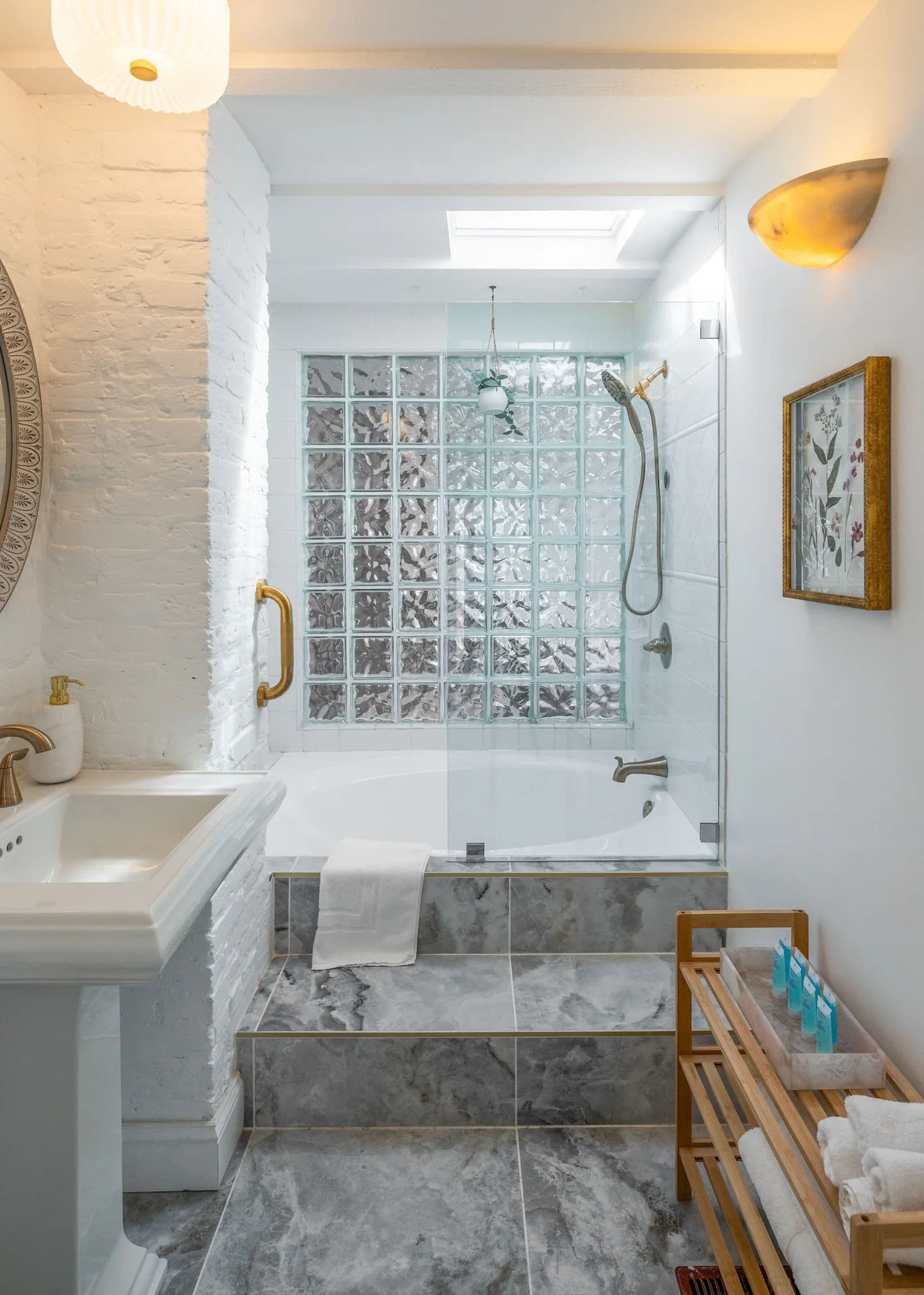 A white bathroom with a pedestal sink and a bathtub with a glass-enclosed shower. A glass block window provides privacy while letting in natural light. A bamboo shelf holds towels and toiletries.