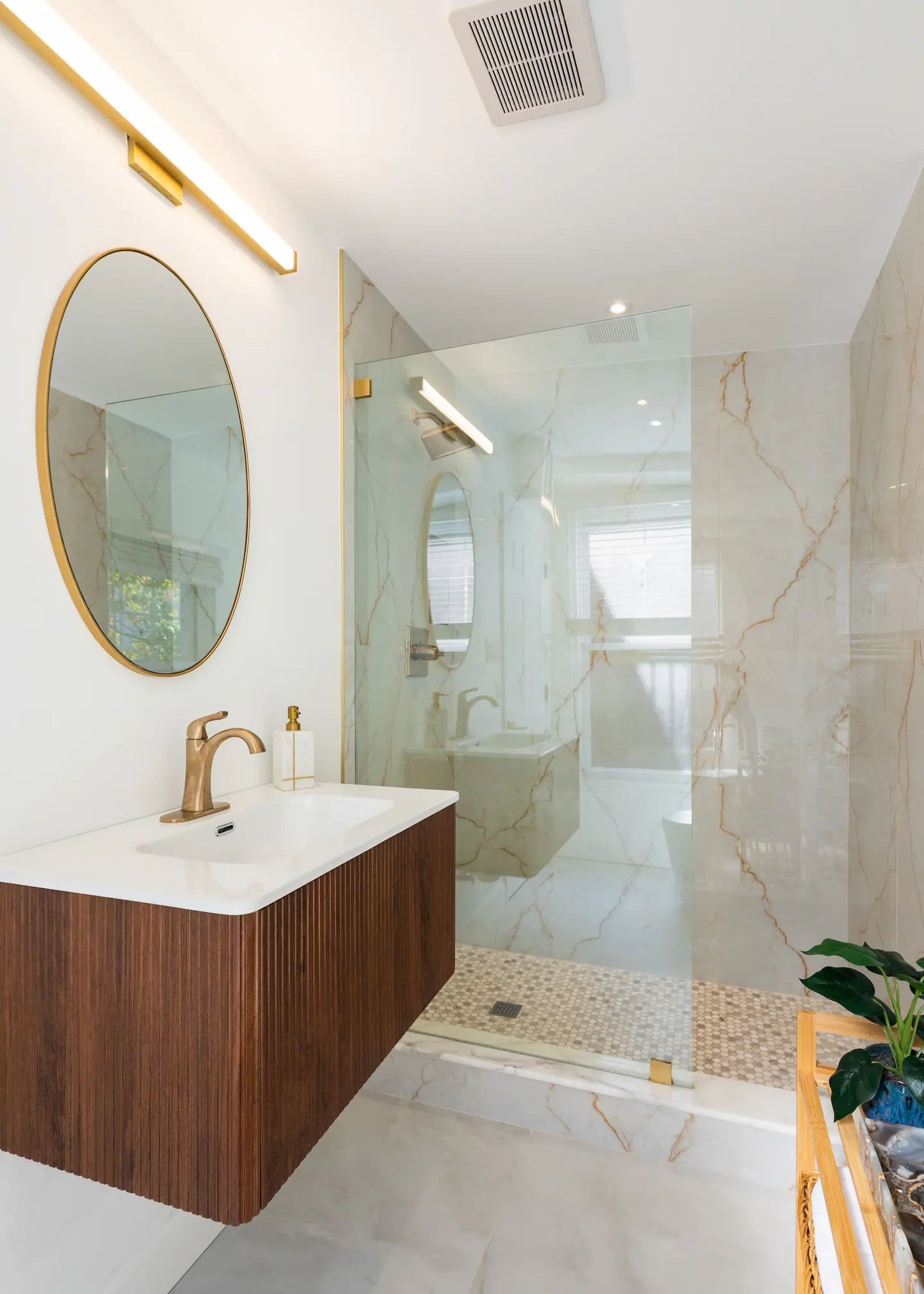 A modern bathroom with a floating wood vanity and a large walk-in shower with a glass door. A round mirror hangs above the sink.