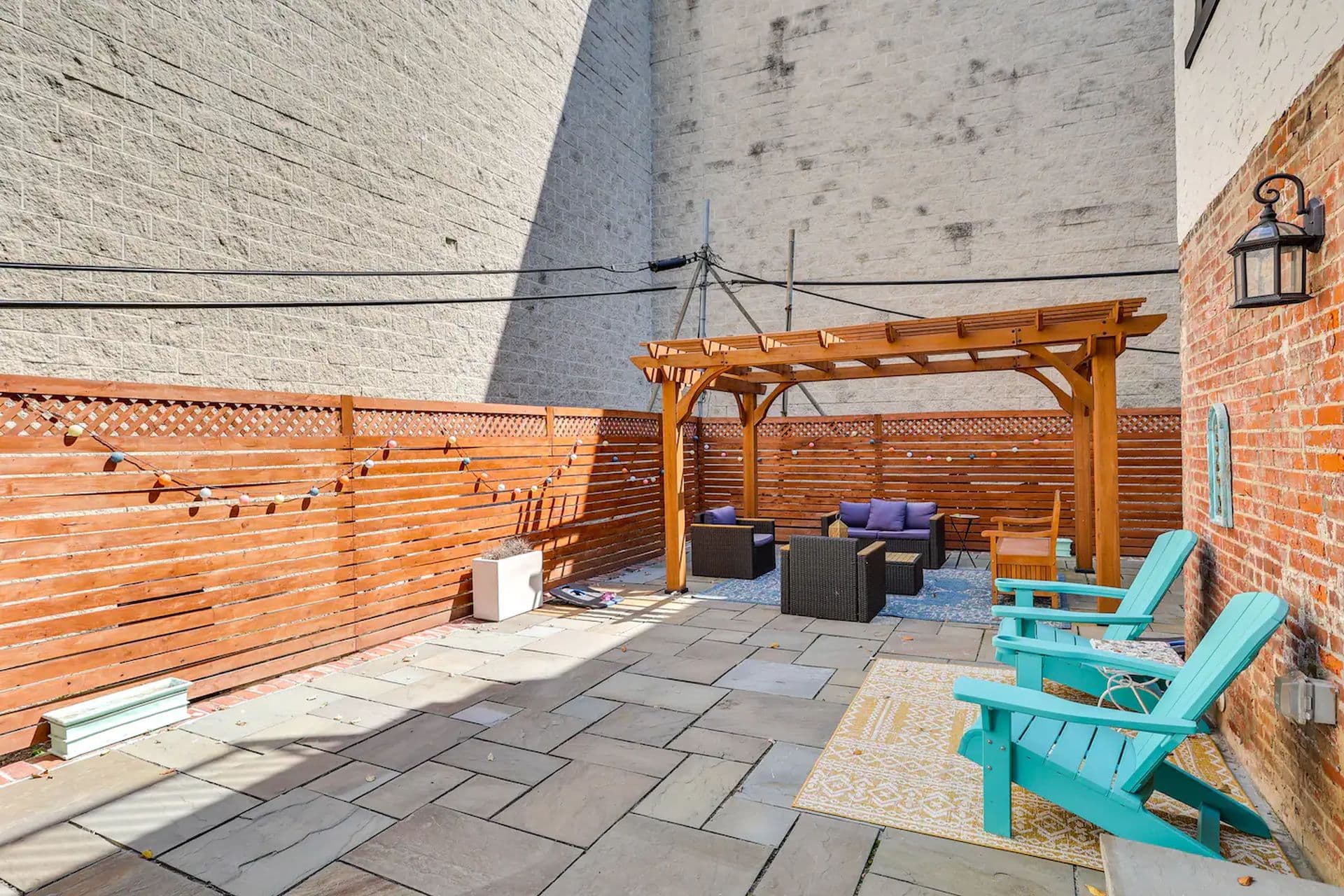 A back patio with a wooden pergola over a seating area with a sofa and chairs. Two turquoise Adirondack chairs sit on the paved patio.
