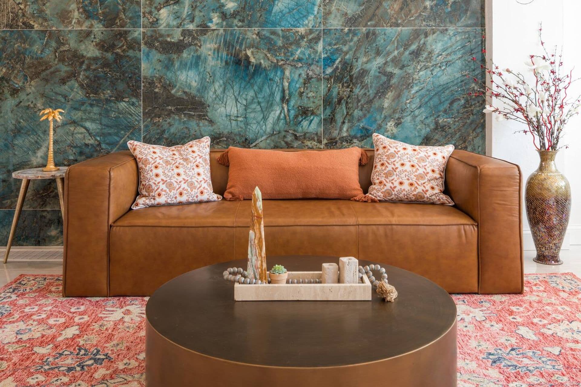 A living room with a brown leather sofa with pillows on a patterned rug. A round coffee table sits in front of the sofa, and a large abstract artwork hangs on the wall.