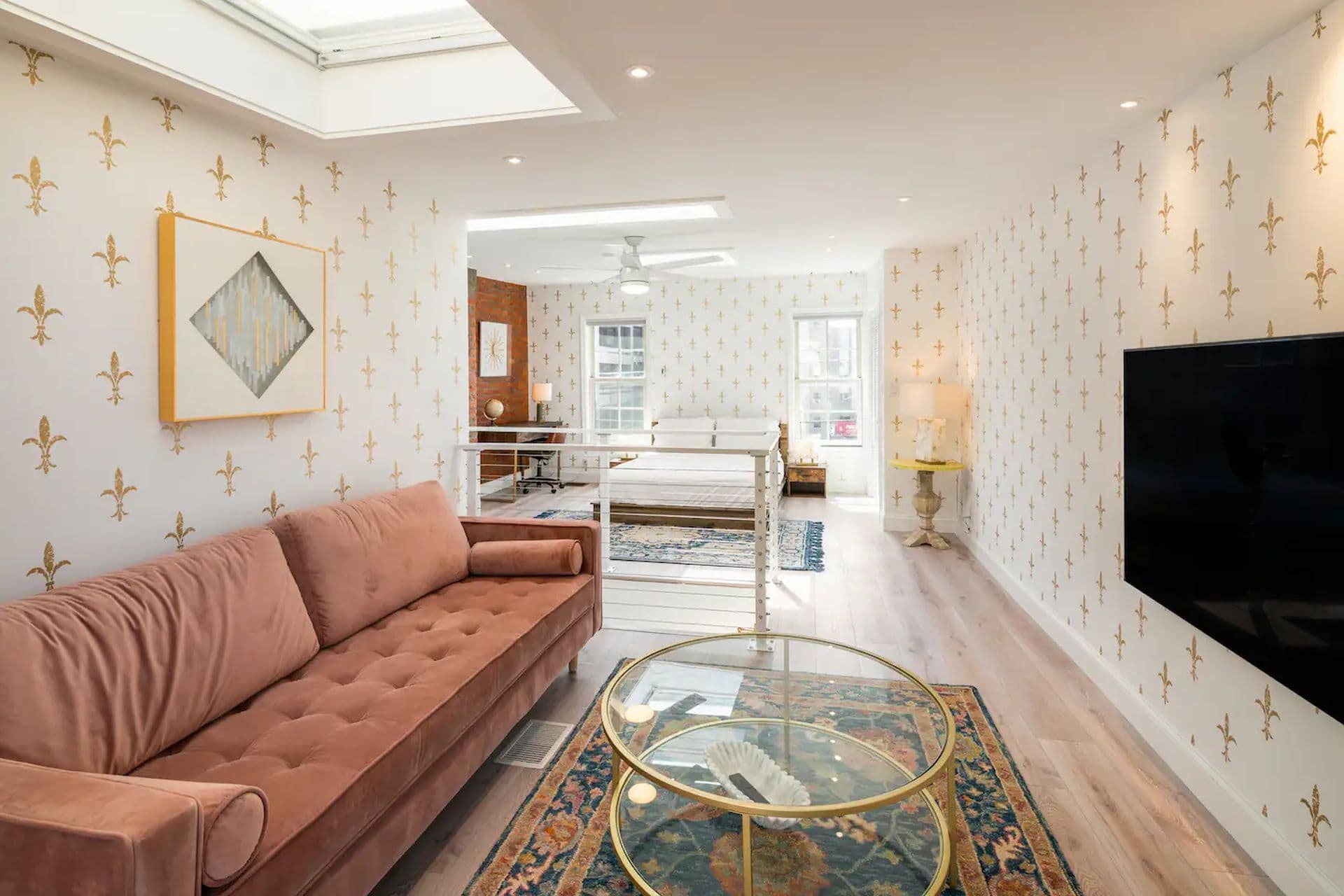 A living room with a pink sofa and a glass coffee table on a patterned rug. A TV is mounted on the wall, and a loft area with a bed is visible in the background.