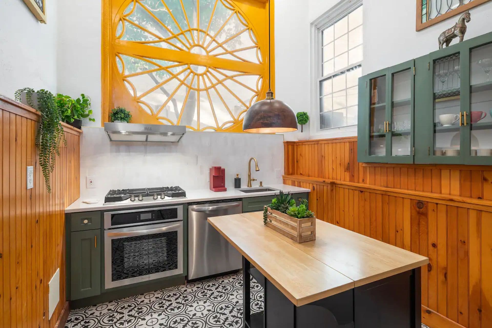 A kitchen with wood paneled walls and a large, round window with a yellow frame. Gray cabinets, a stainless steel oven, and a wooden island with a plant create a modern rustic feel. Black and white patterned floor tiles complete the look.