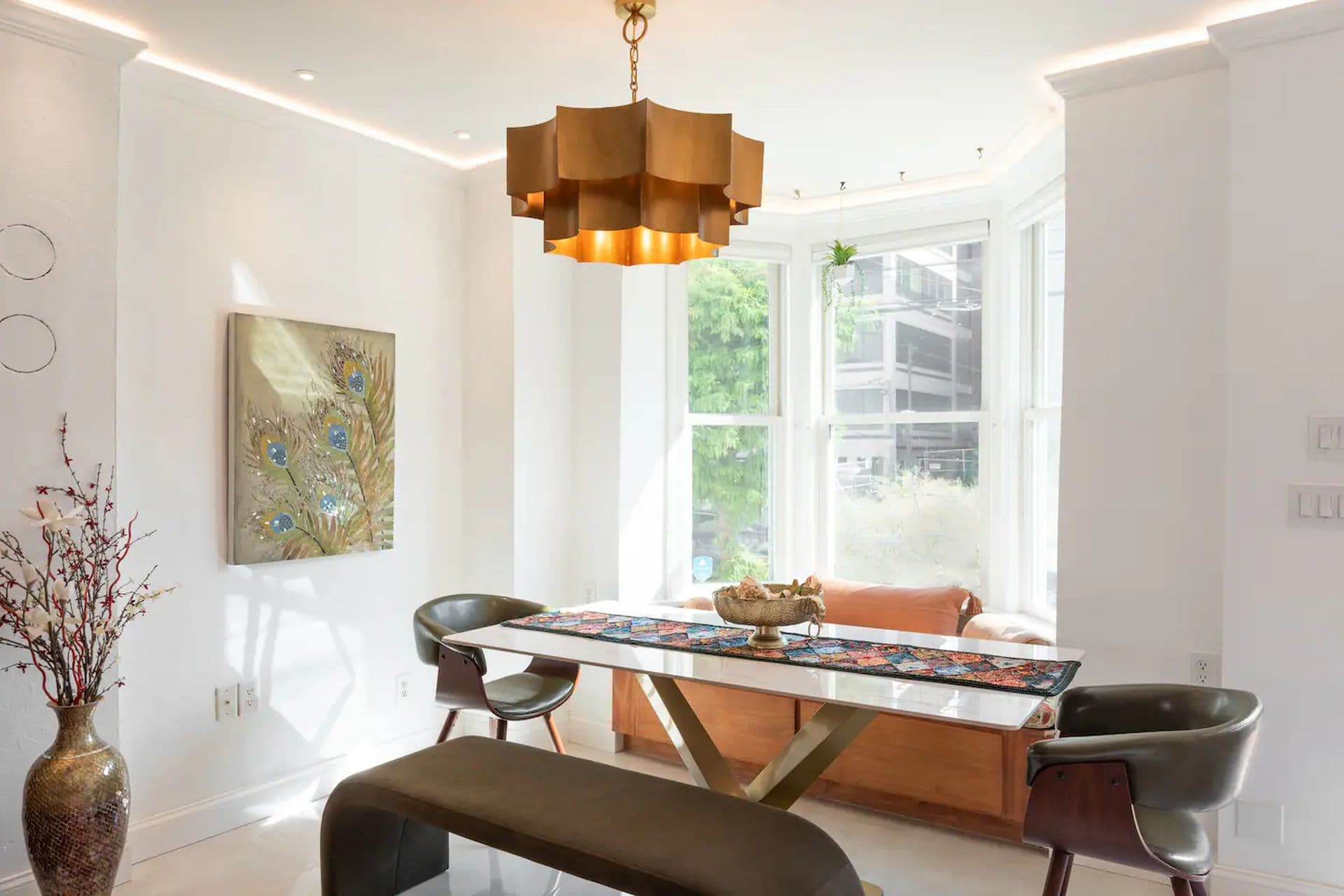 A bright dining room with a large window and a gold geometric light fixture. A long table with place settings is surrounded by chairs, and a painting hangs on the wall.