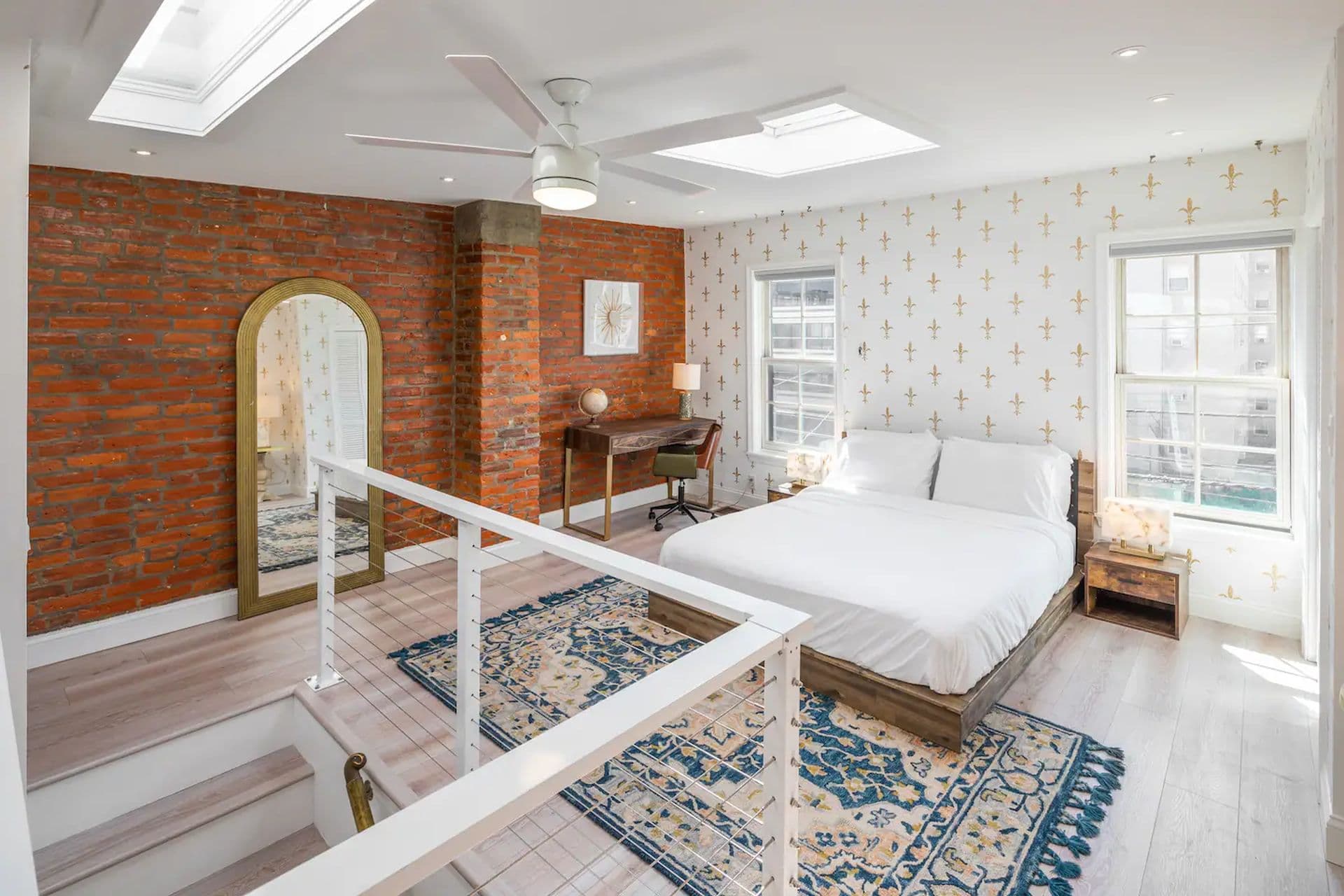 A bedroom with a king-size bed and a floral wallpaper on one wall. A brick wall with a large mirror is on the opposite side. A small desk sits by the window, and a skylight provides natural light.