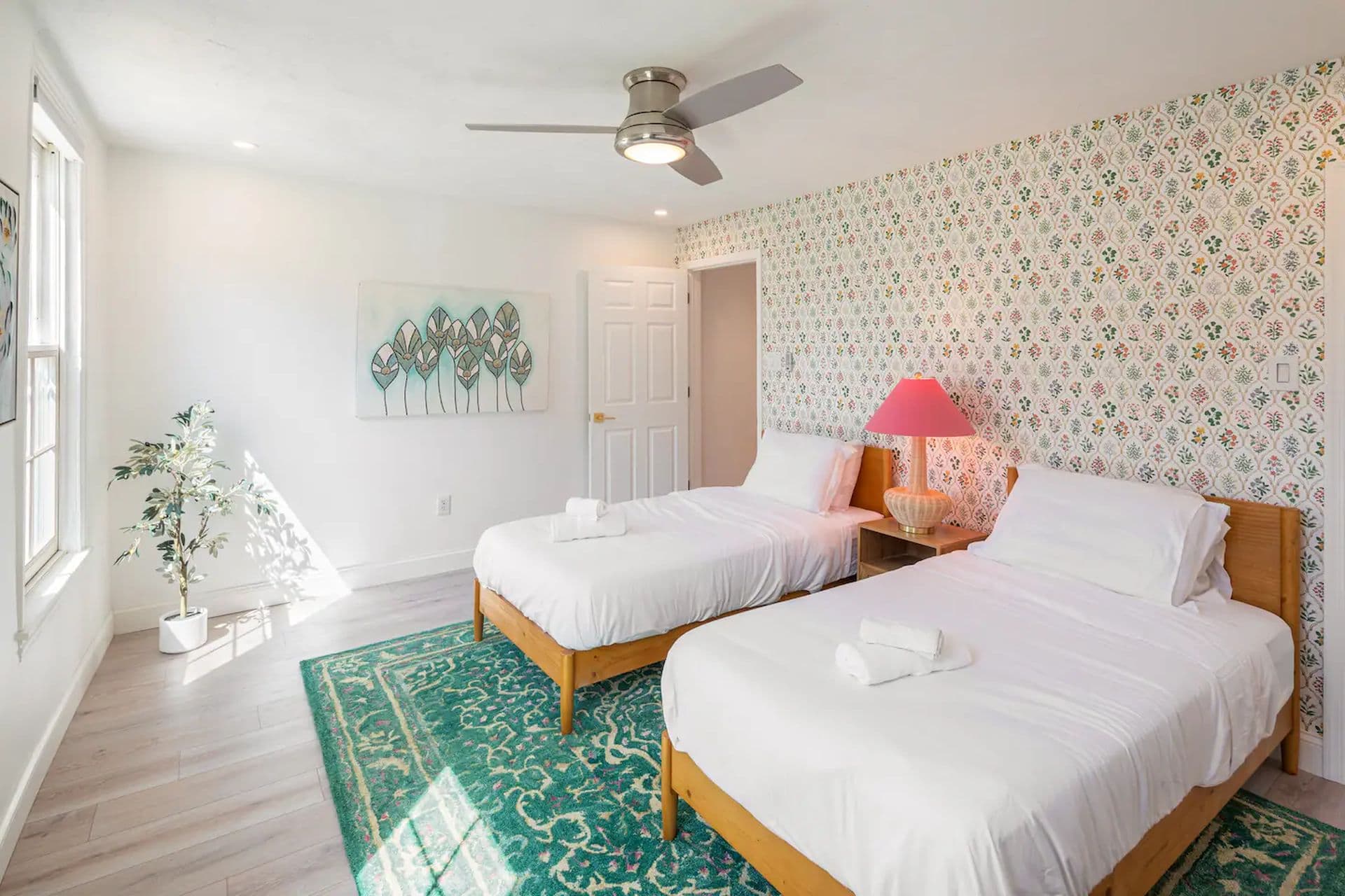 A bedroom with two single beds with white comforters on a green rug. A patterned wallpaper covers one wall, and a window is on the opposite wall.