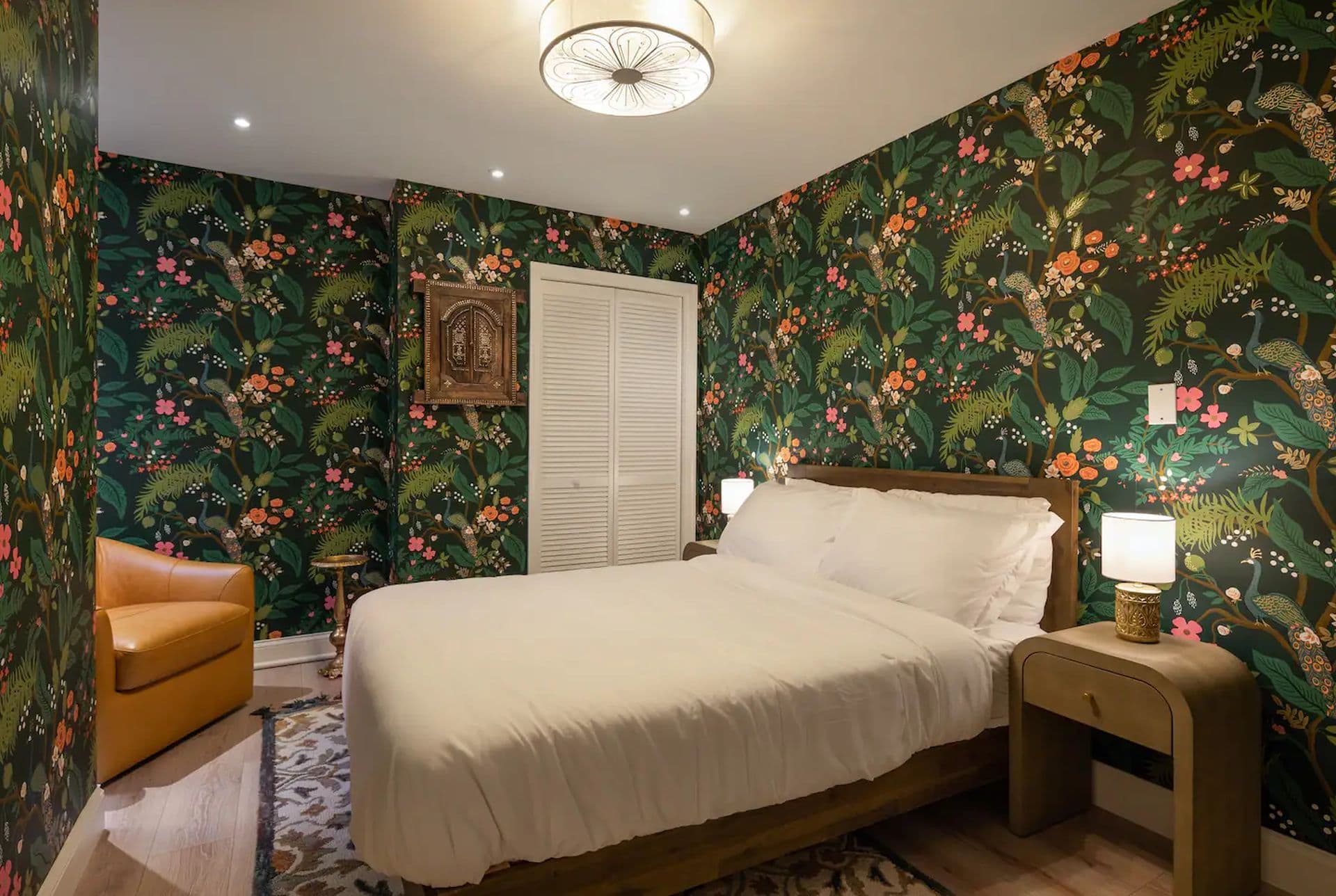 A bedroom with a queen-size bed and a tropical-themed wallpaper featuring lush green foliage and exotic birds. A leather armchair sits in the corner, and a small table with a lamp is beside the bed.