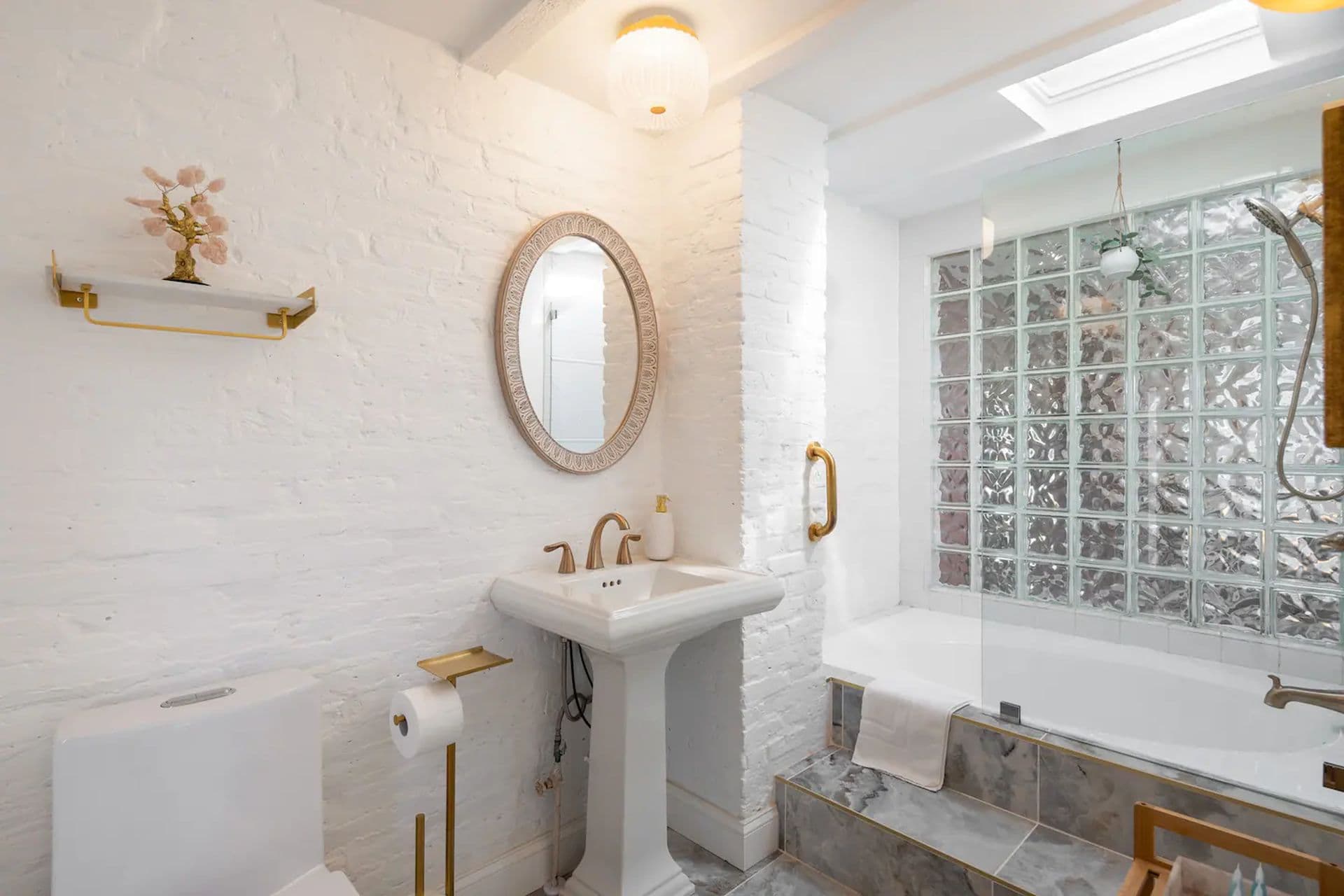 A white bathroom with a pedestal sink and a bathtub with a glass block window. A decorative shelf hangs above the sink.
