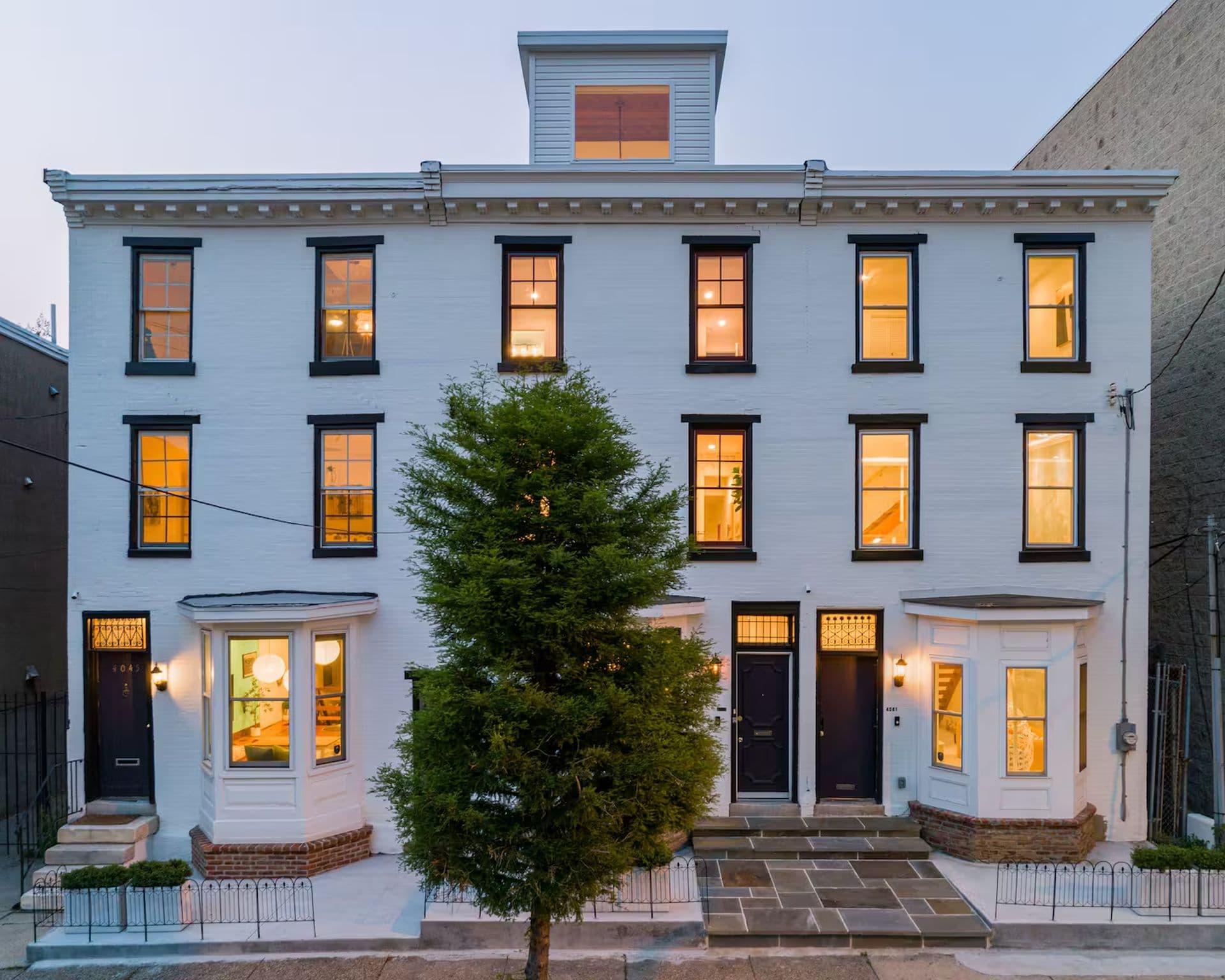 The front exterior of a white three-story row house with black trim and evenly spaced windows. A small tree stands in front of the building.