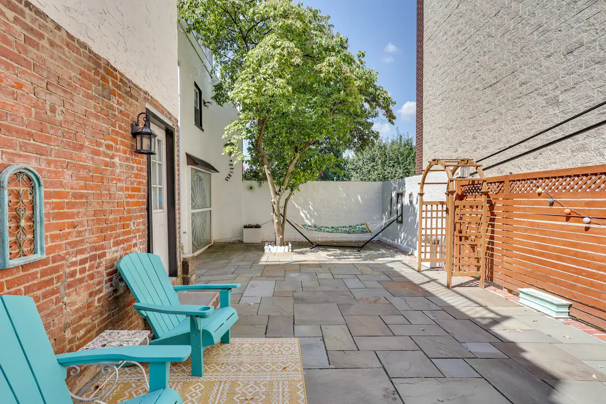 A paved backyard patio with two turquoise Adirondack chairs and a hammock strung between a tree and a brick wall.