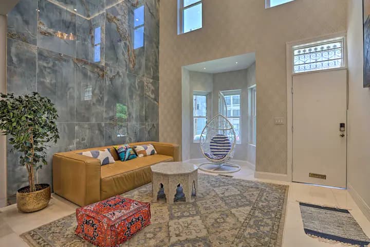 A modern living room with a high ceiling and a brown sofa on a patterned rug. A large textured wall provides a dramatic backdrop. A glimpse of the entryway is visible in the background.