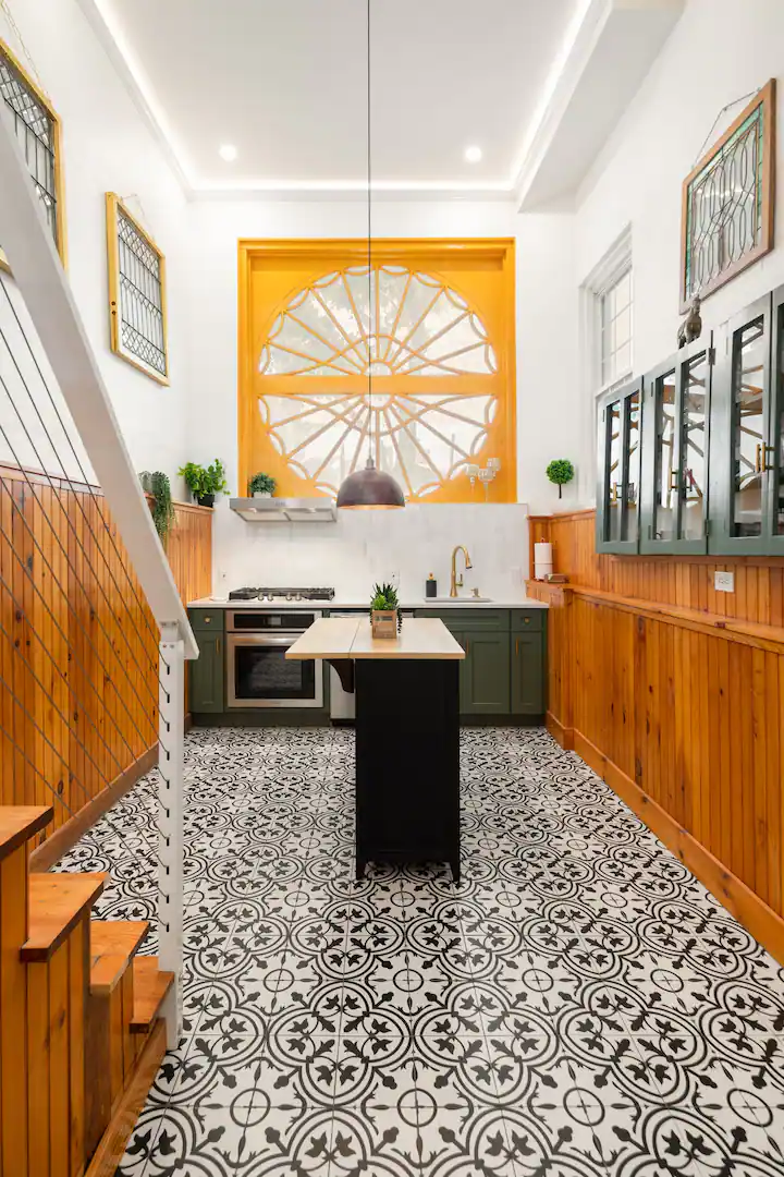 A galley kitchen with wood-paneled walls and a large, round window with a yellow frame. Dark gray cabinets, a stainless steel oven, and a black island with a plant create a modern rustic feel. Black and white patterned floor tiles complete the look. A wooden staircase is on the left.