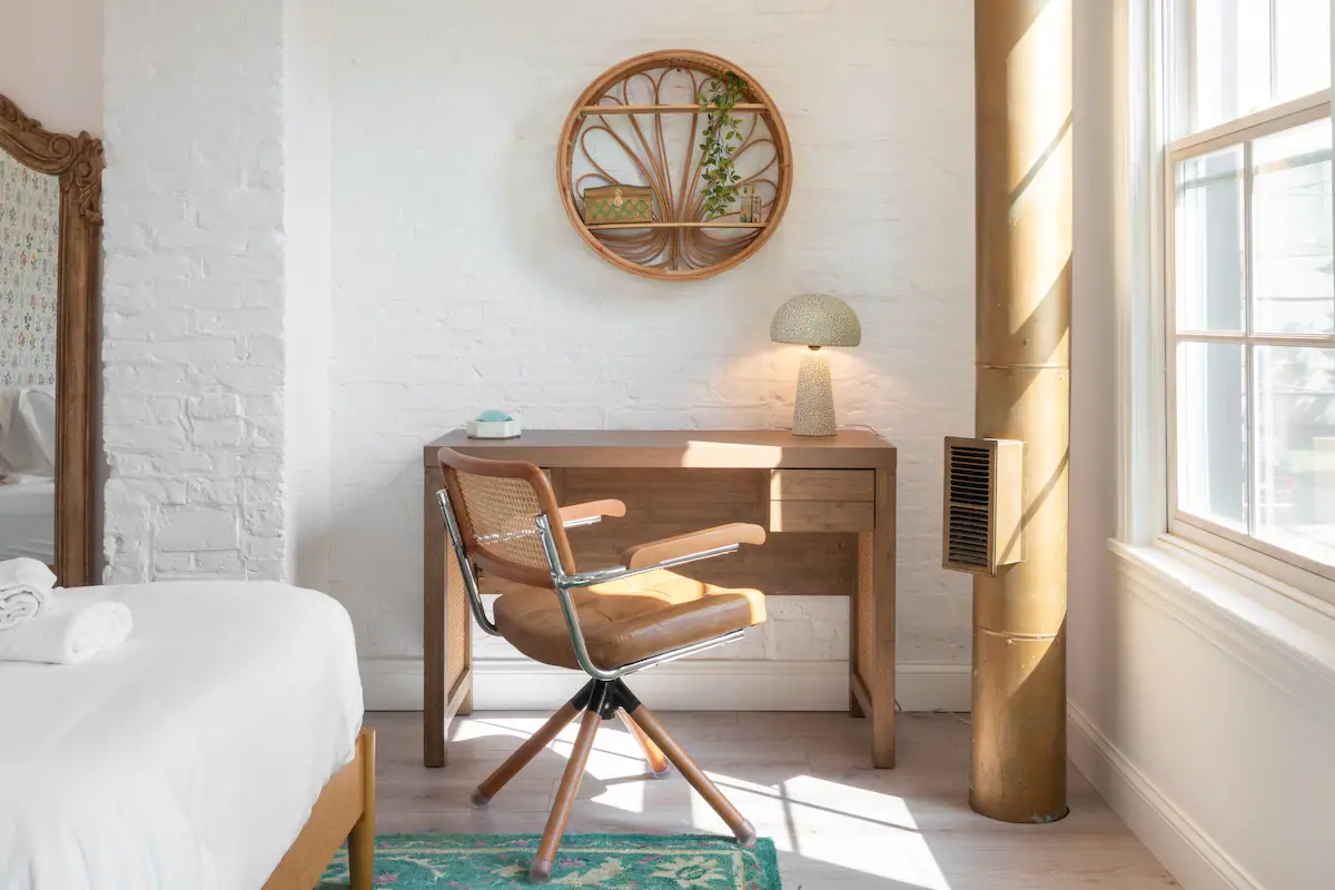 A corner of a bedroom with a wooden desk and chair, a round mirror on the wall, and a window letting in natural light.