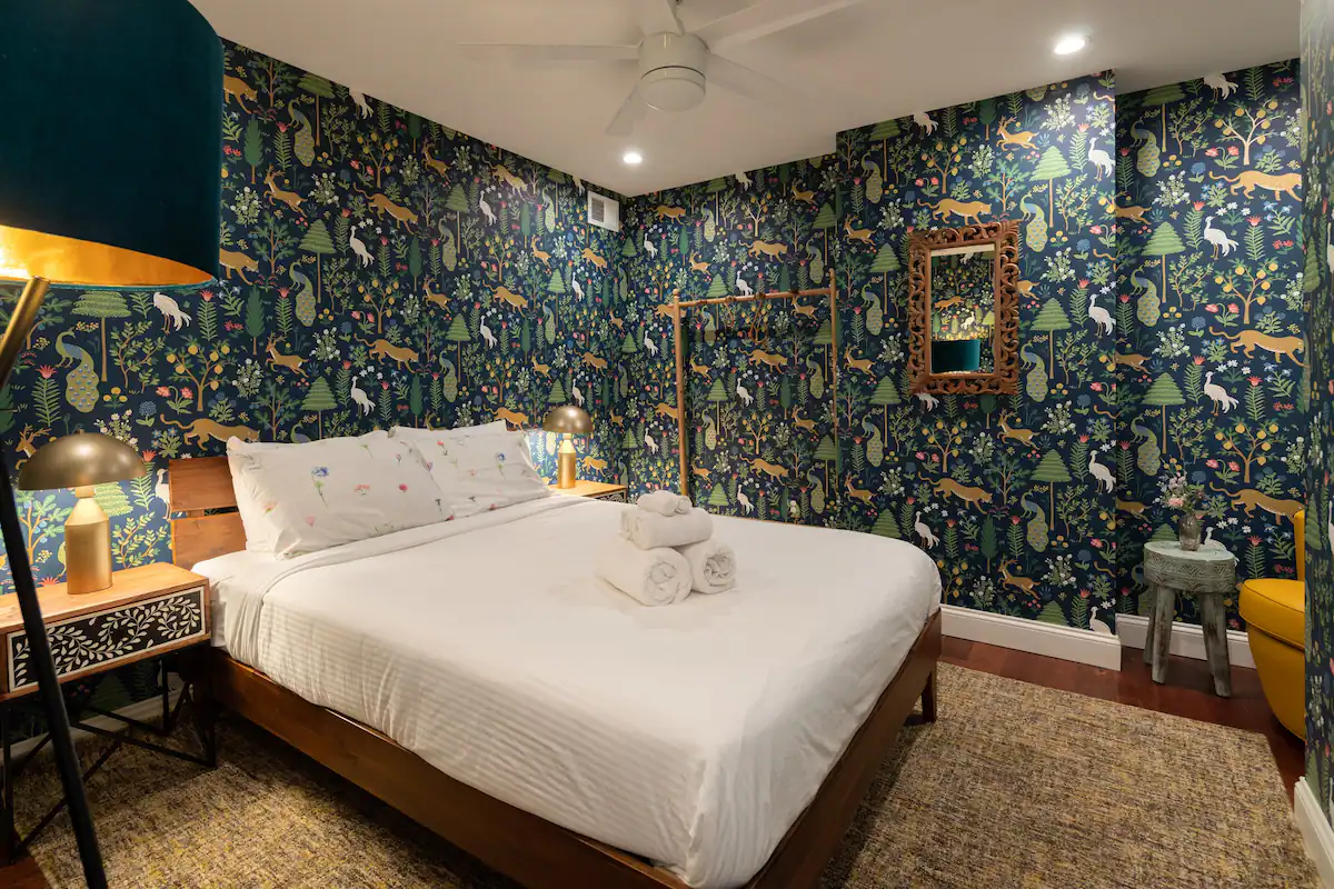 A cozy bedroom with a queen-size bed and a dark green patterned wallpaper featuring birds and plants. A floor lamp stands beside the bed, and a small table with a mirror is on the opposite wall.