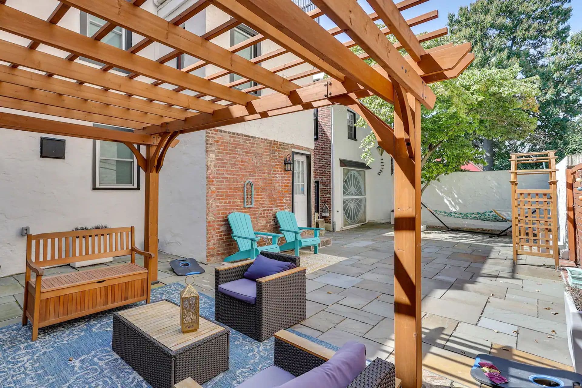 A backyard patio area with a wooden pergola providing shade. Two wicker chairs with blue cushions sit on an outdoor rug, facing a small table. A hammock is strung across the back of the yard.