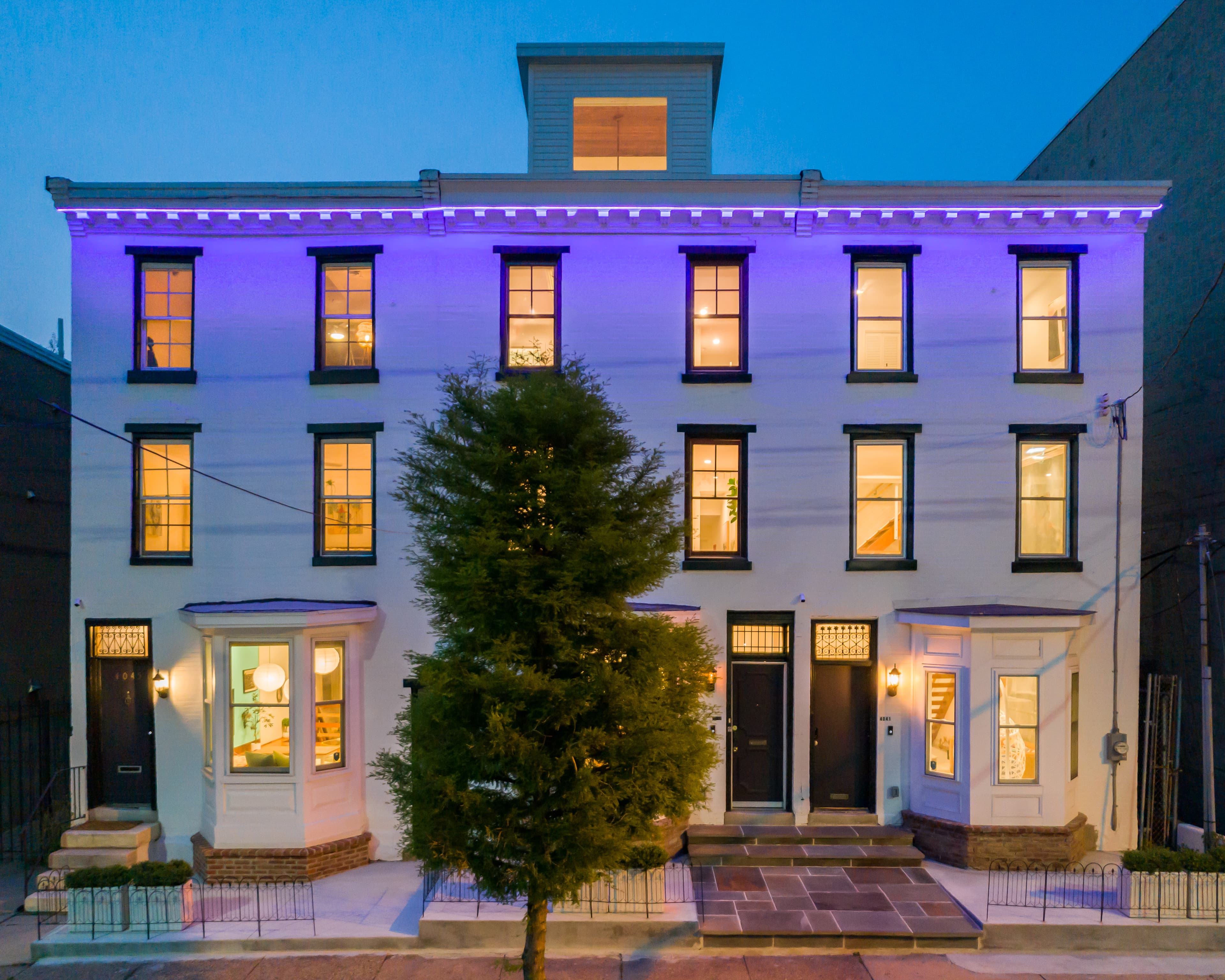 A modern, three-story white townhouse illuminated with purple lighting at dusk.