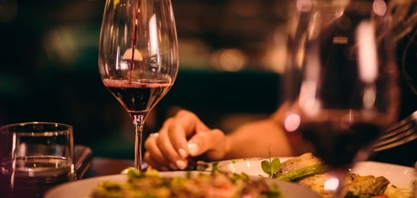 A hand reaches for a plate of food beside a glass of red wine in a dimly lit setting. A hand reaches for a plate of food beside a glass of red wine in a dimly lit setting.