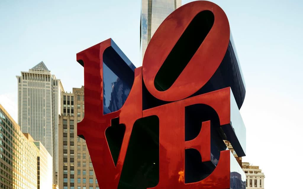 A large, colorful "LOVE" sculpture stands in an urban setting with skyscrapers in the background.