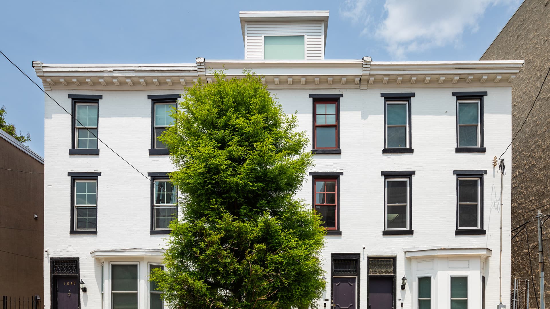 A white three-story building with multiple windows and a large tree in front.