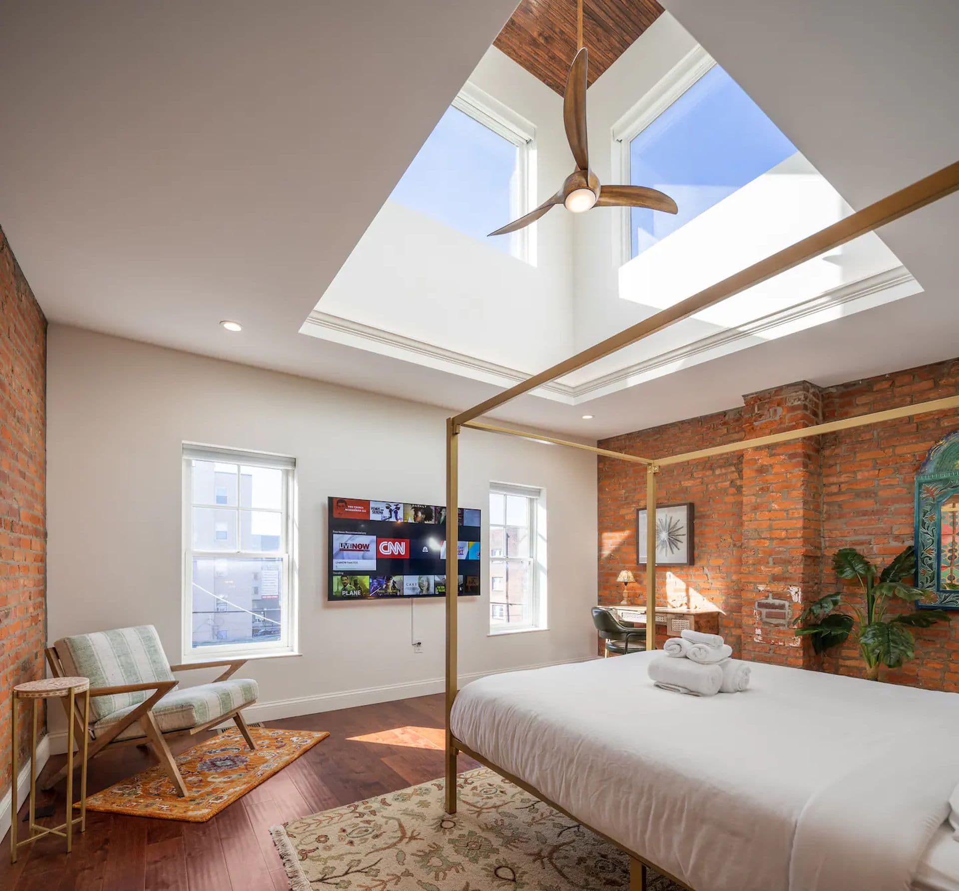 A bedroom with a four-poster bed and a skylight overhead. A brick wall and wood floors add a rustic touch. A TV is mounted on the wall and an armchair sits in the corner.