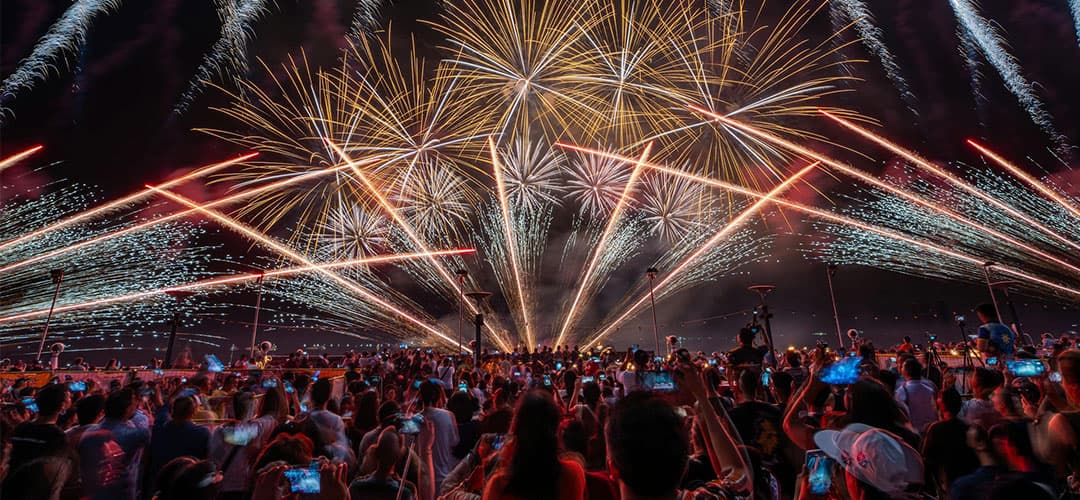 A vibrant display of fireworks illuminates the night sky above a crowd holding smartphones.