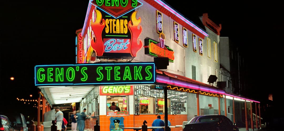Brightly lit exterior of Geno's Steaks restaurant at night, showcasing neon signs and patrons.